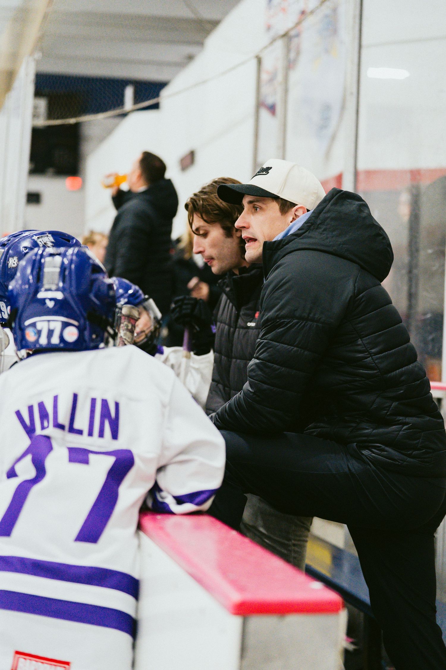 Coaches and players gather at hockey bench during game with player number 77 visible.