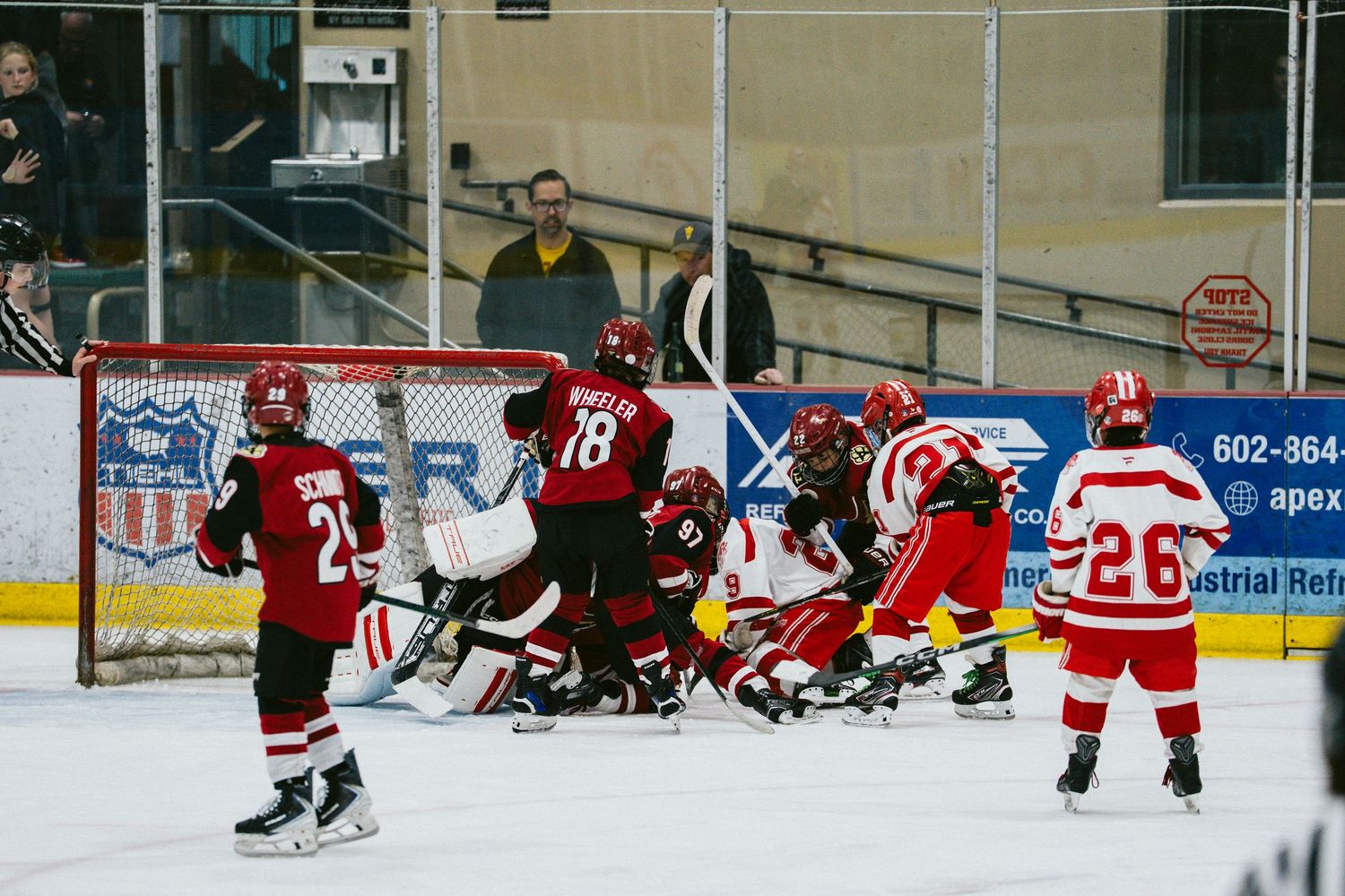Multiple hockey players scramble for the puck during an intense goal-mouth action at an indoor rink.