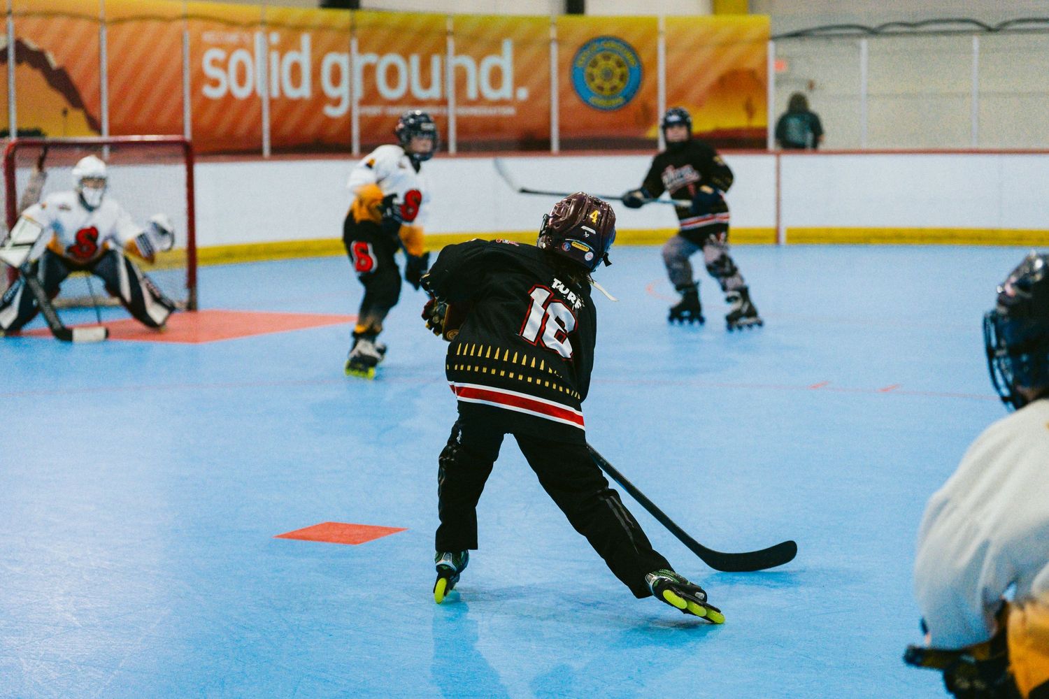 Youth roller hockey player in black jersey winds up for a shot during an indoor rink game.
