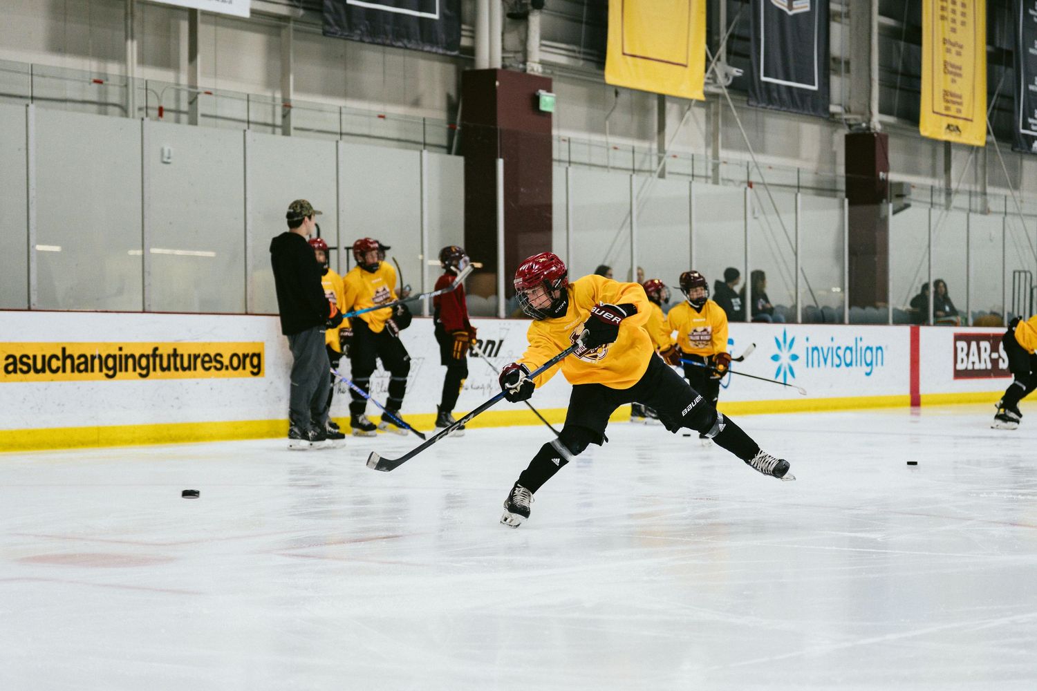 Youth hockey players in yellow jerseys take shots on goal during an indoor ice hockey training session.