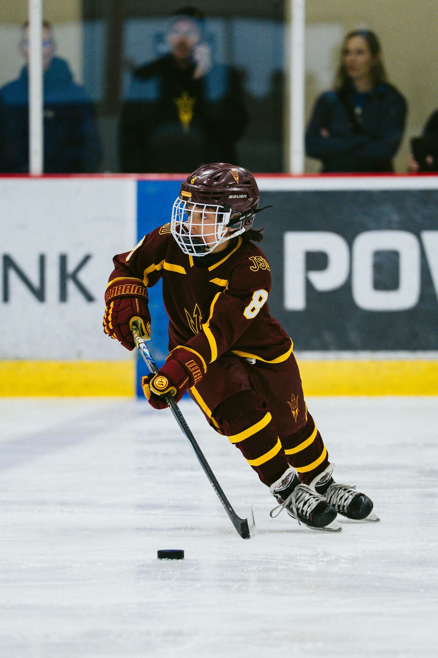 Youth hockey player in brown and gold jersey skating with puck on ice rink during game.