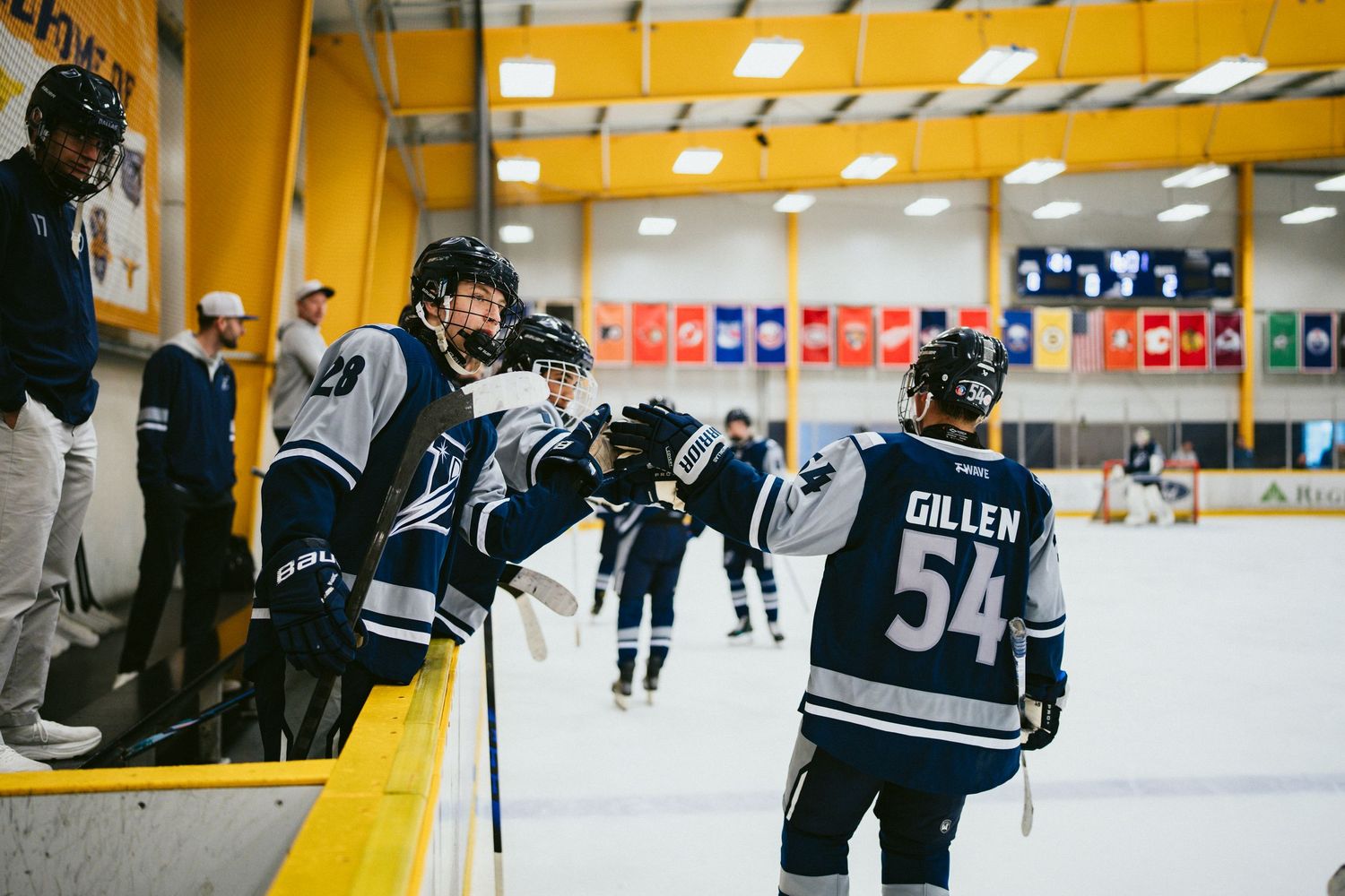 Hockey players in blue and white uniforms gather at the bench during an indoor ice hockey game, player Gillen wearing number 54.