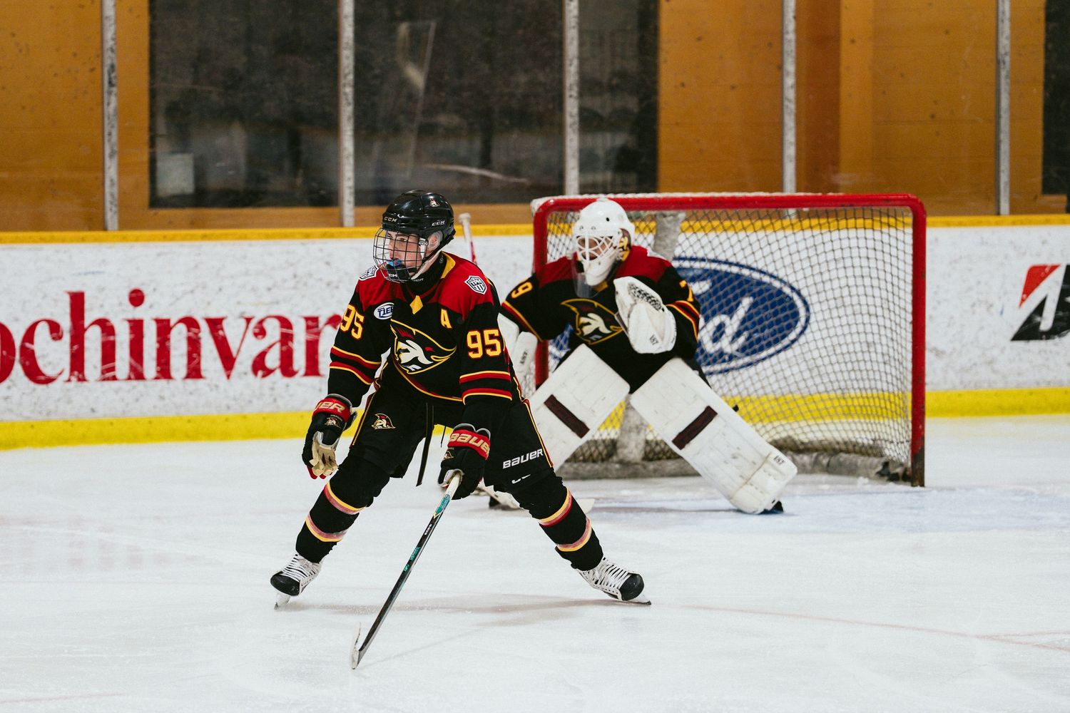 Ice hockey player in black and red jersey controls puck near goal during competitive game on indoor rink.