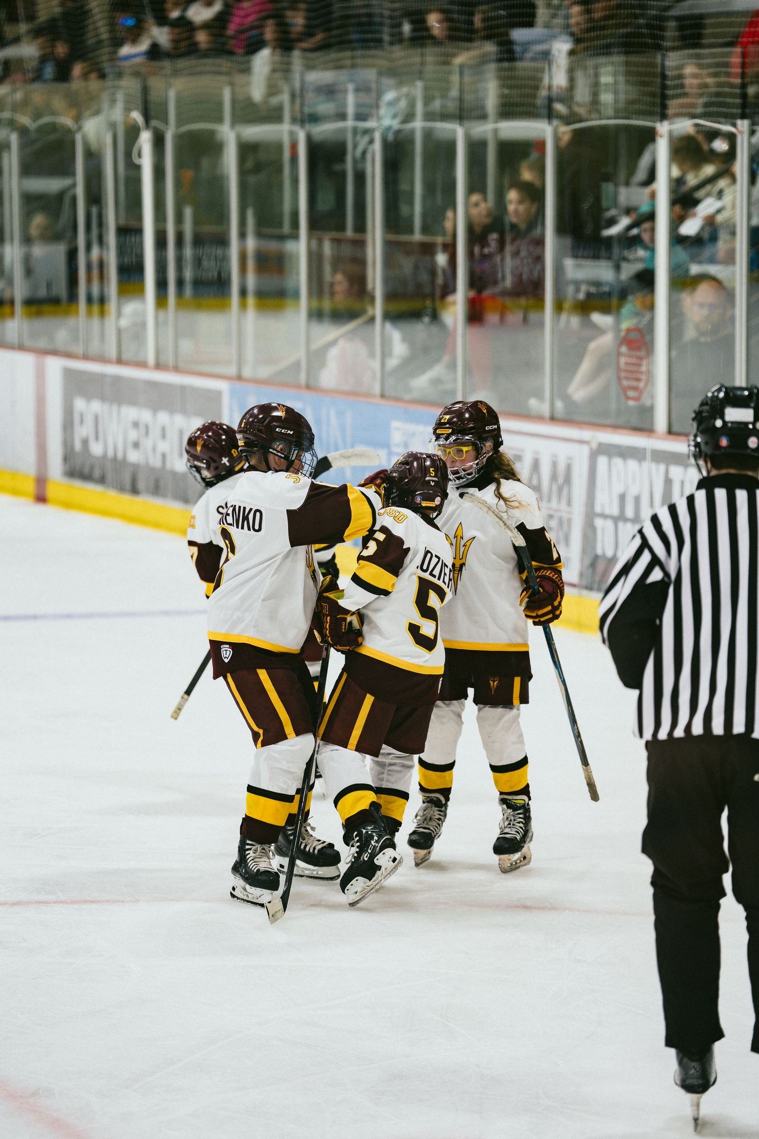 Youth ice hockey players in maroon and gold celebrate a goal together on the ice near a referee.
