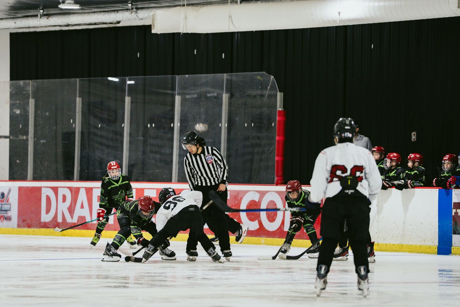 Referee oversees a face-off between youth hockey teams in black and red uniforms on indoor ice rink.
