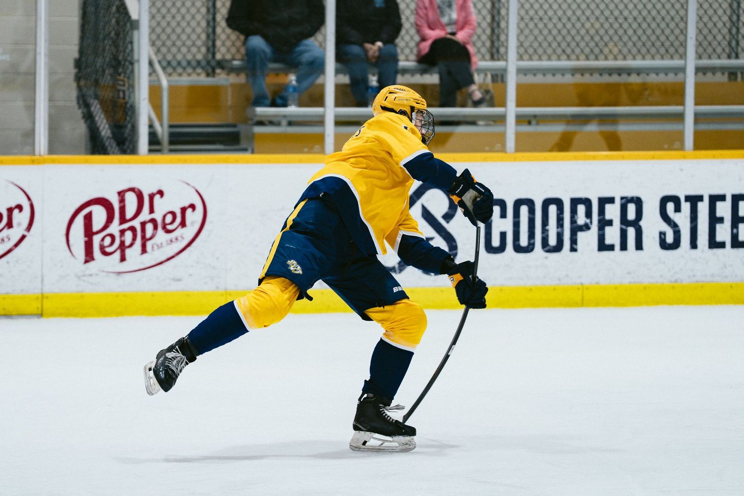 Youth hockey player in yellow uniform takes a powerful slap shot on ice in front of a Dr Pepper banner.