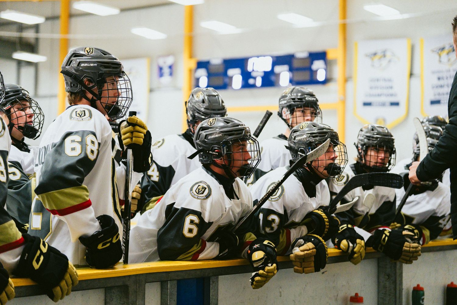Youth ice hockey players in black and gold uniforms crowd the bench during a game.