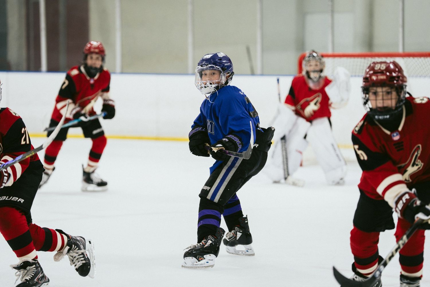 Youth hockey player in blue jersey skates among opponents in red during competitive ice hockey game.