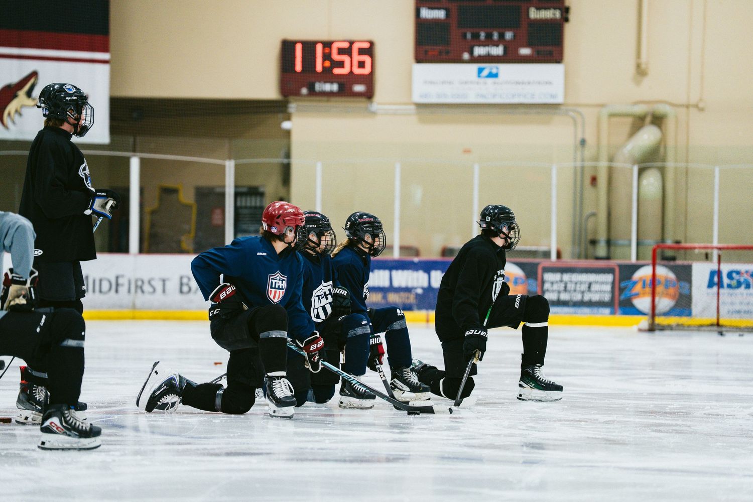 Group of young hockey players kneel on ice during team practice with scoreboard showing 11:56.