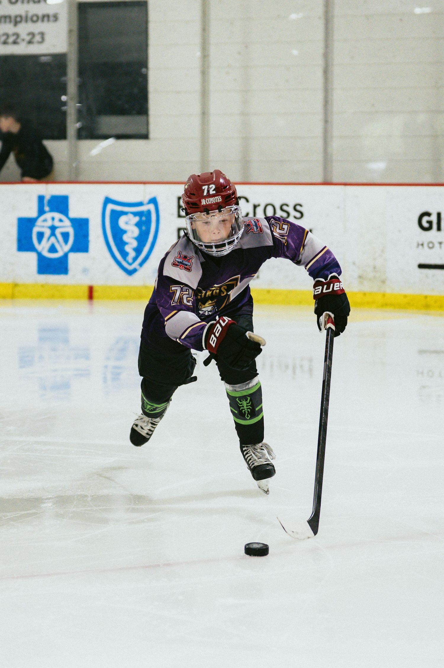 Youth hockey player number 72 in black and purple uniform controls puck on indoor ice rink.