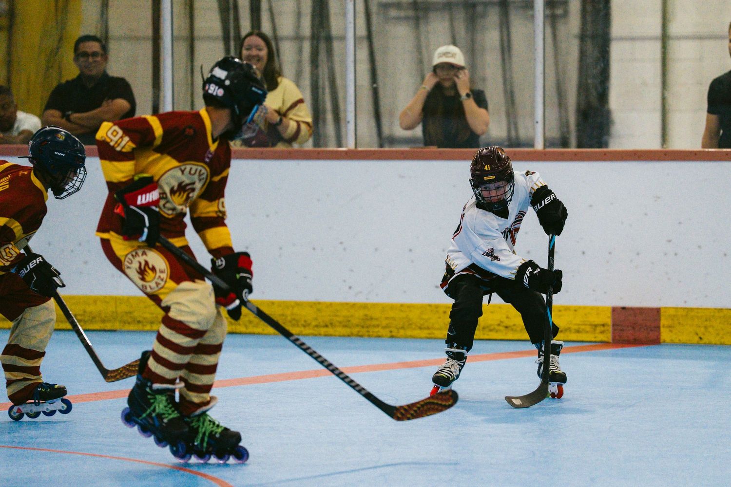 Players compete in an intense inline roller hockey game on an indoor rink, wearing team jerseys and wielding hockey sticks.