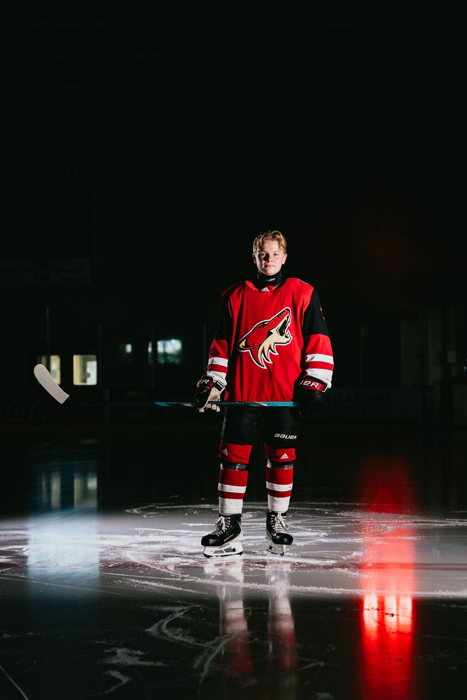 Ice hockey player standing on rink in red Arizona Coyotes jersey holding stick under dramatic lighting.