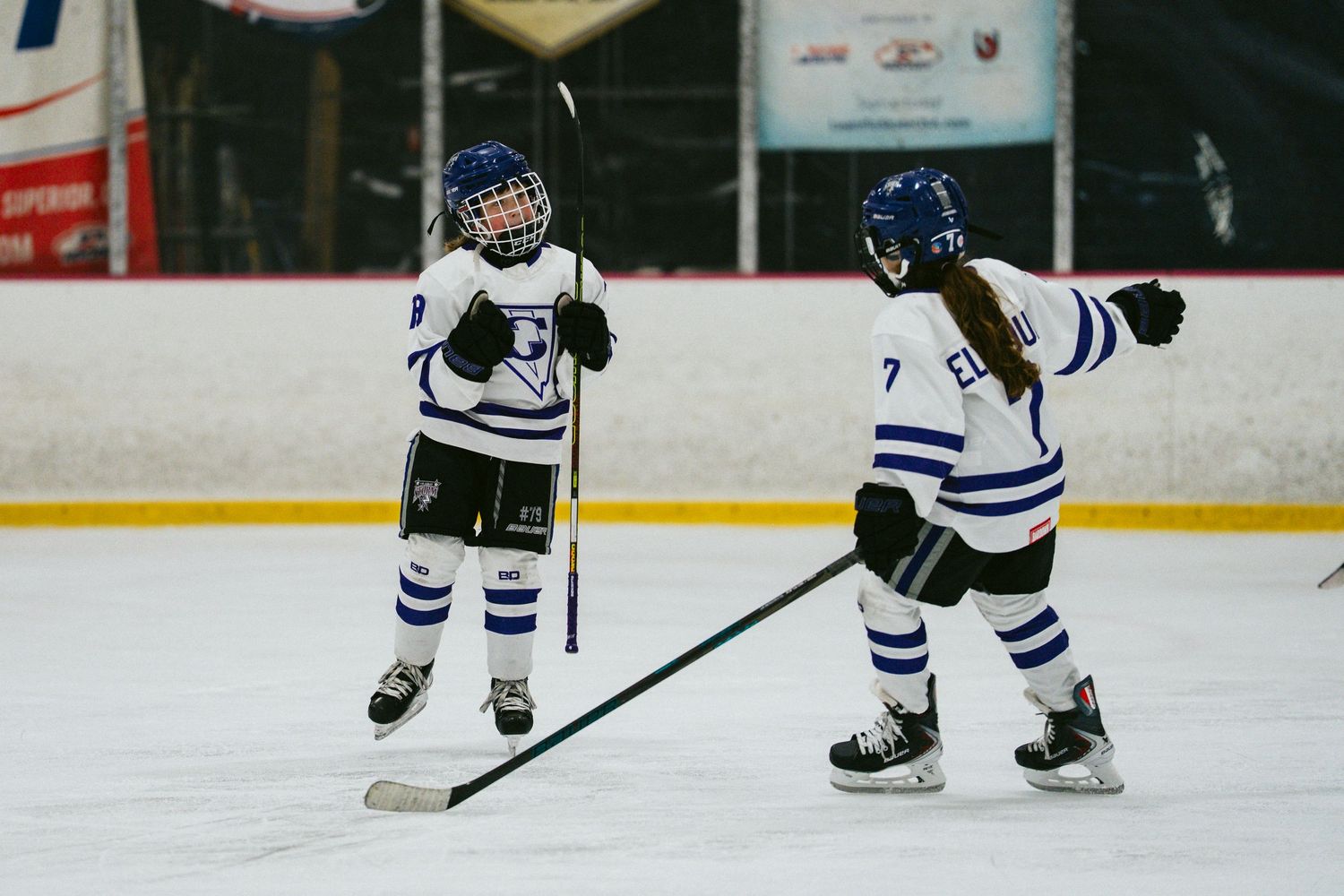 Two young hockey players in matching uniforms skate on ice during game.