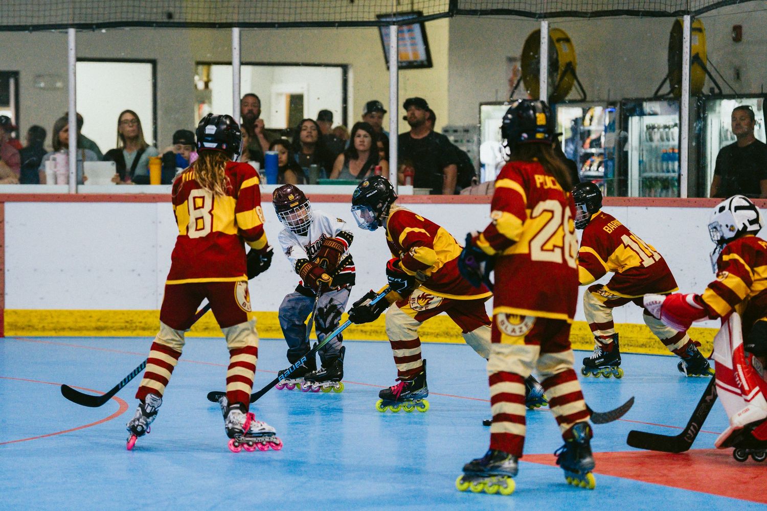 Roller hockey players in red and yellow uniforms compete on an indoor rink with spectators watching from the stands.