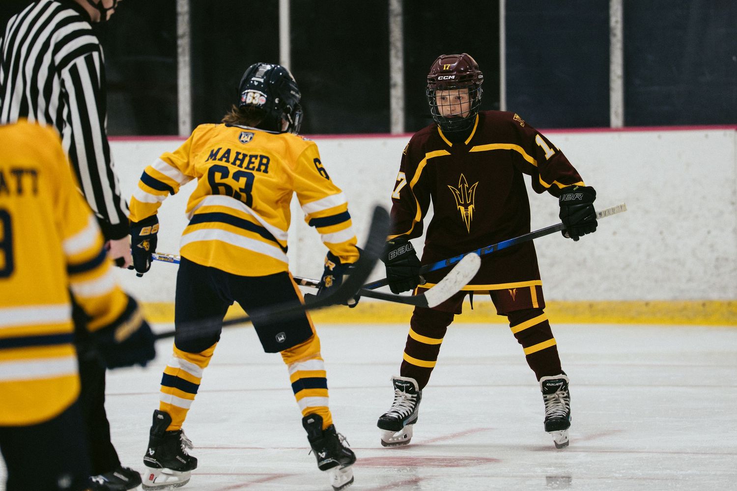 Two youth hockey players face off on the ice during a game, wearing yellow and maroon uniforms.