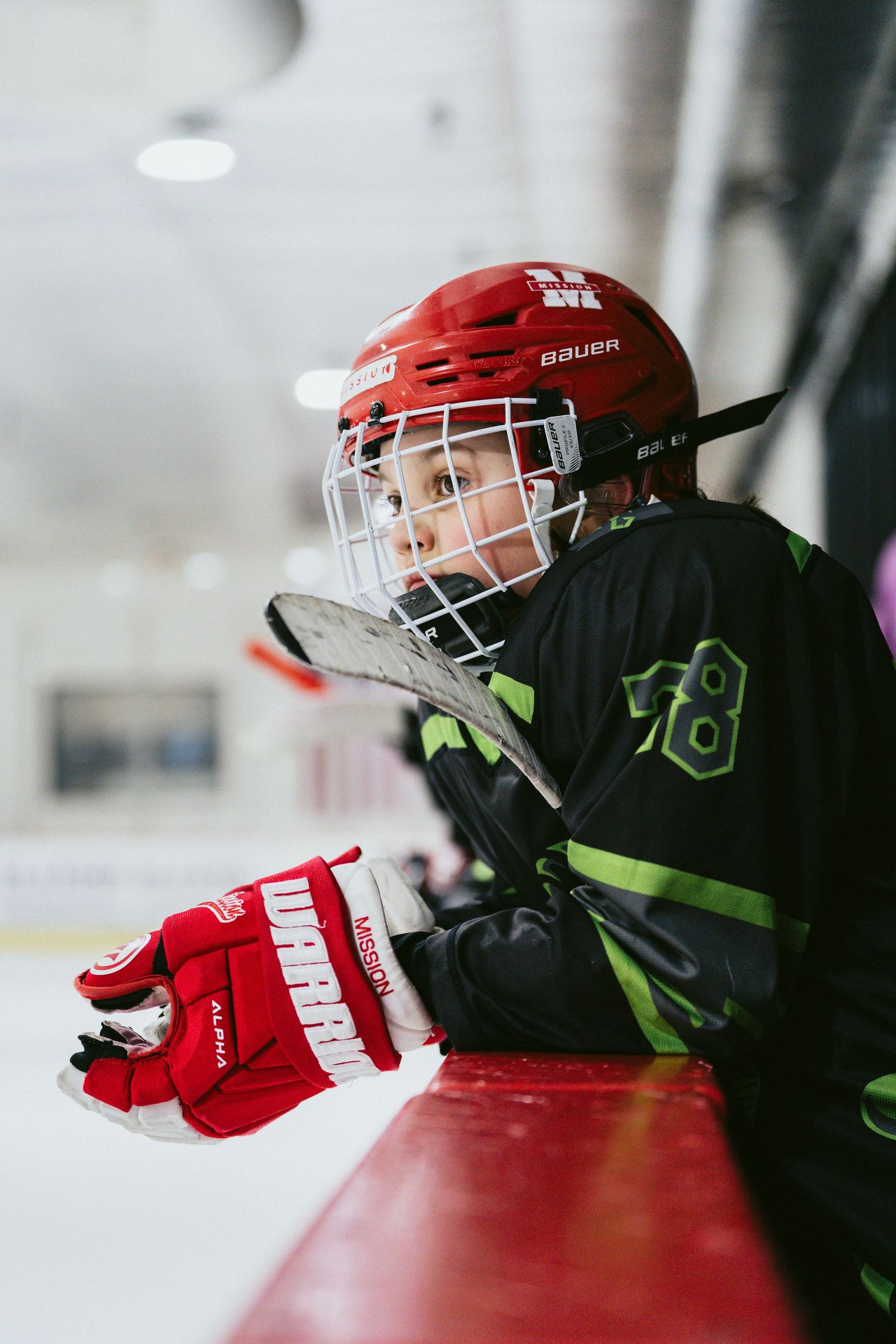Young hockey player in red Bauer helmet leaning on boards watching youth ice hockey game.