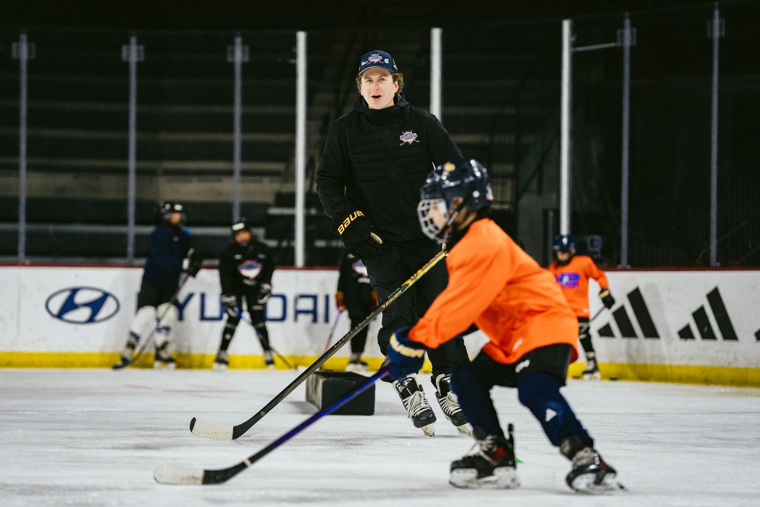 Hockey coach instructing young players in orange and black jerseys during an indoor ice hockey training drill.