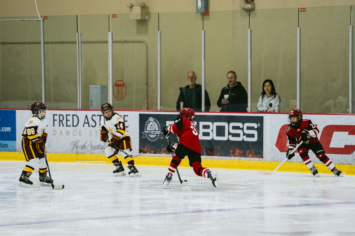 Hockey players in red and black jerseys practicing drills on an ice rink with advertisements on the boards.