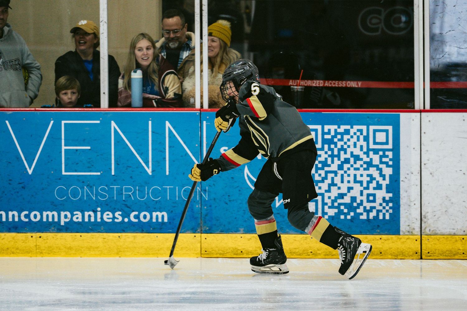 Hockey player in teal jersey skating with the puck past advertising boards.