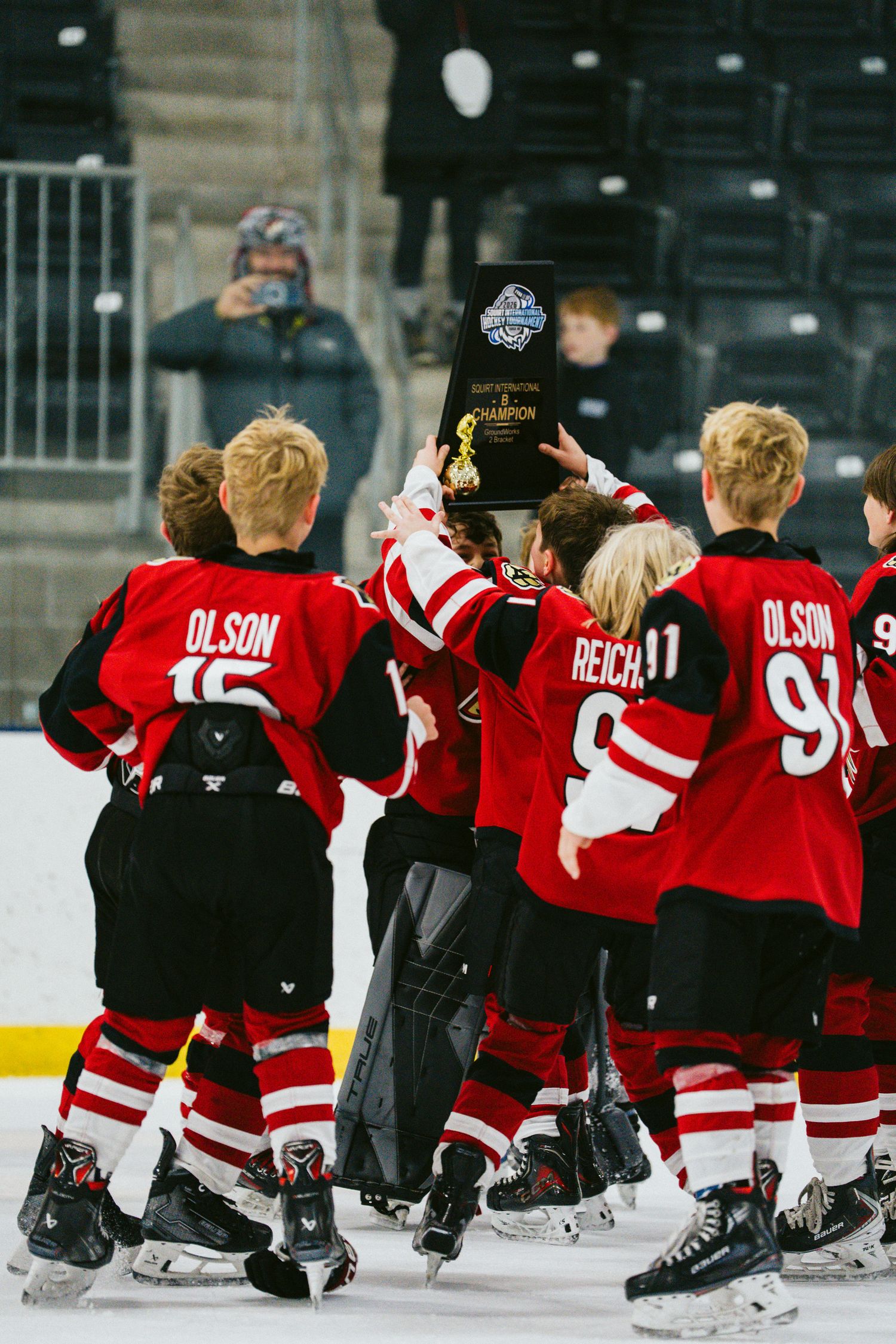 Hockey players in red jerseys sitting in formation on ice rink during team activity.