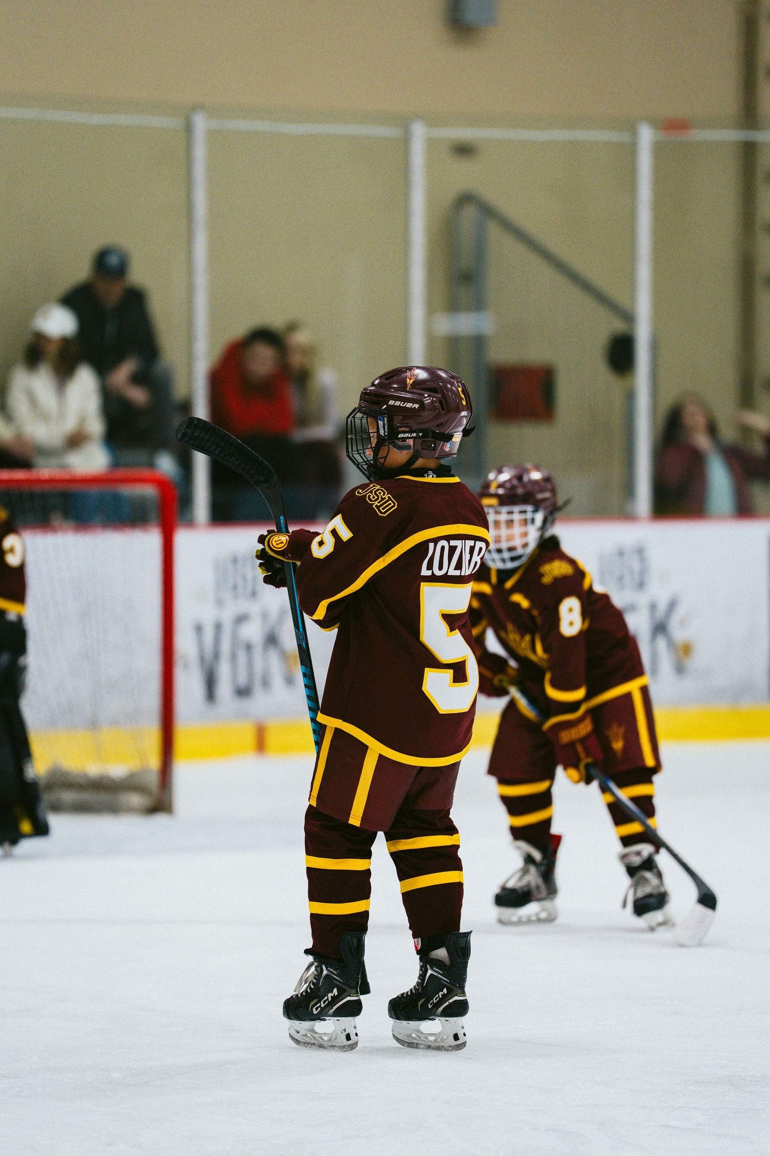 Young hockey player in brown and yellow uniform stands on ice during youth hockey game with teammates and spectators.