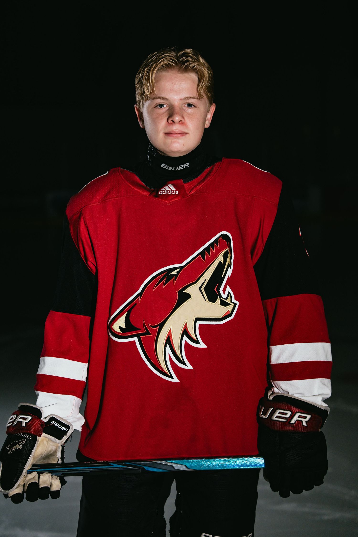 Young hockey player in Arizona Coyotes red jersey holding a Bauer stick, posing on dark ice rink background.