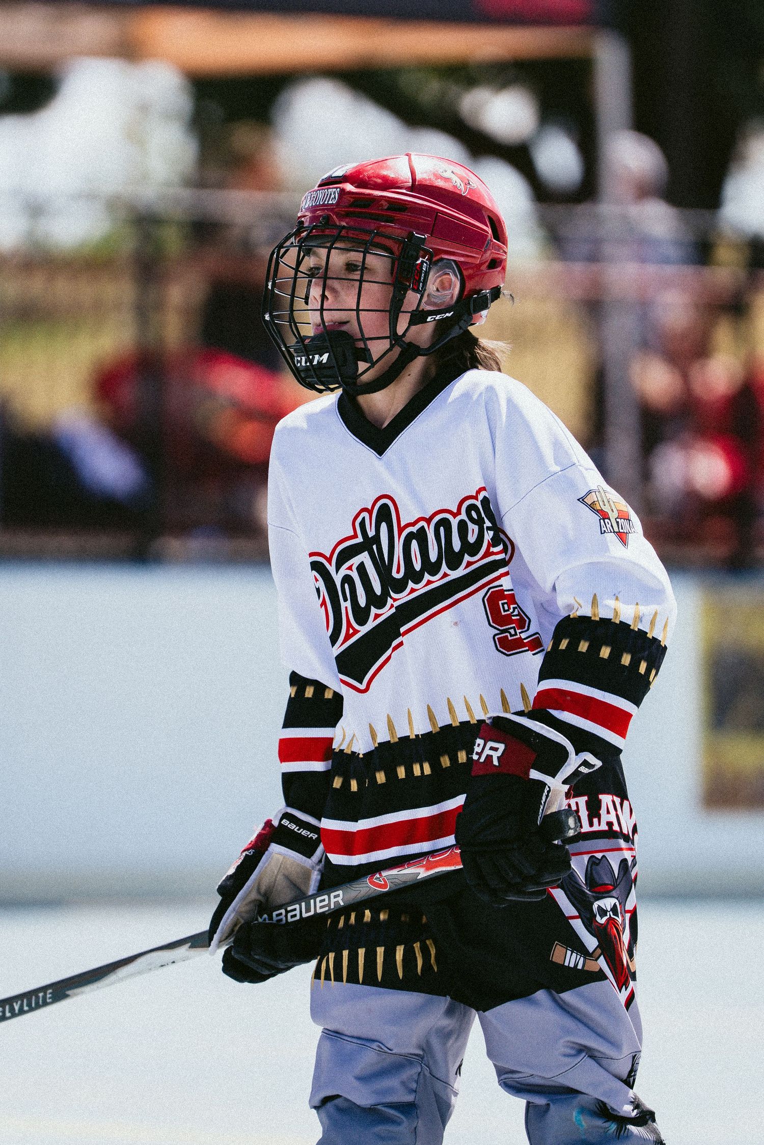 Young roller hockey player in white Outlaws jersey and red helmet holds a stick during an outdoor rink game.