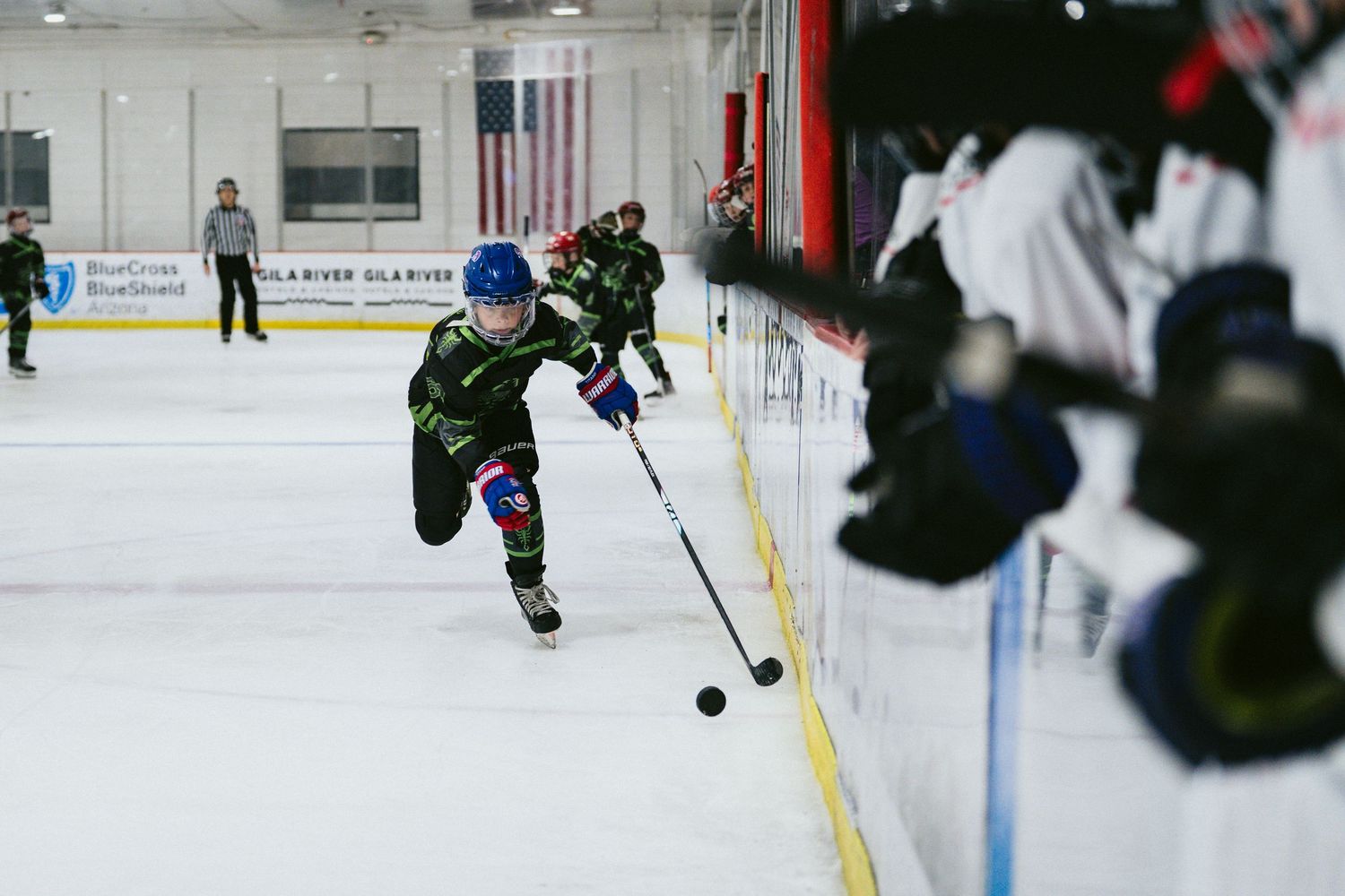 Young hockey player in black green uniform chases puck along boards during youth ice hockey game.