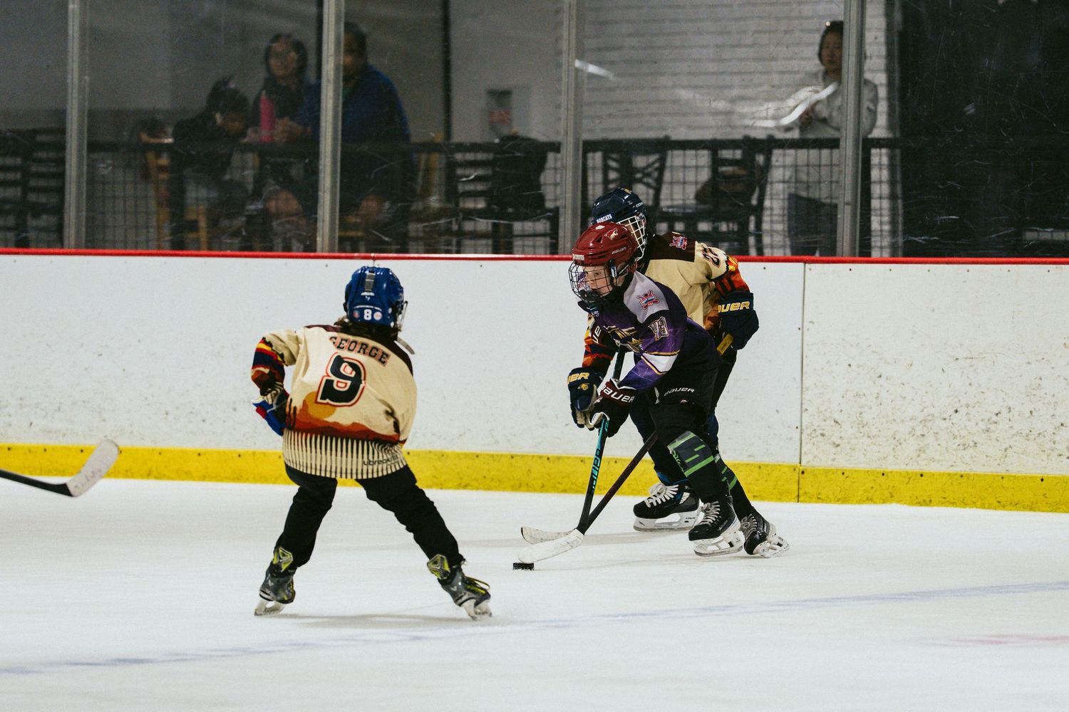 Two youth hockey players in gold and white uniforms compete for the puck on indoor ice rink.