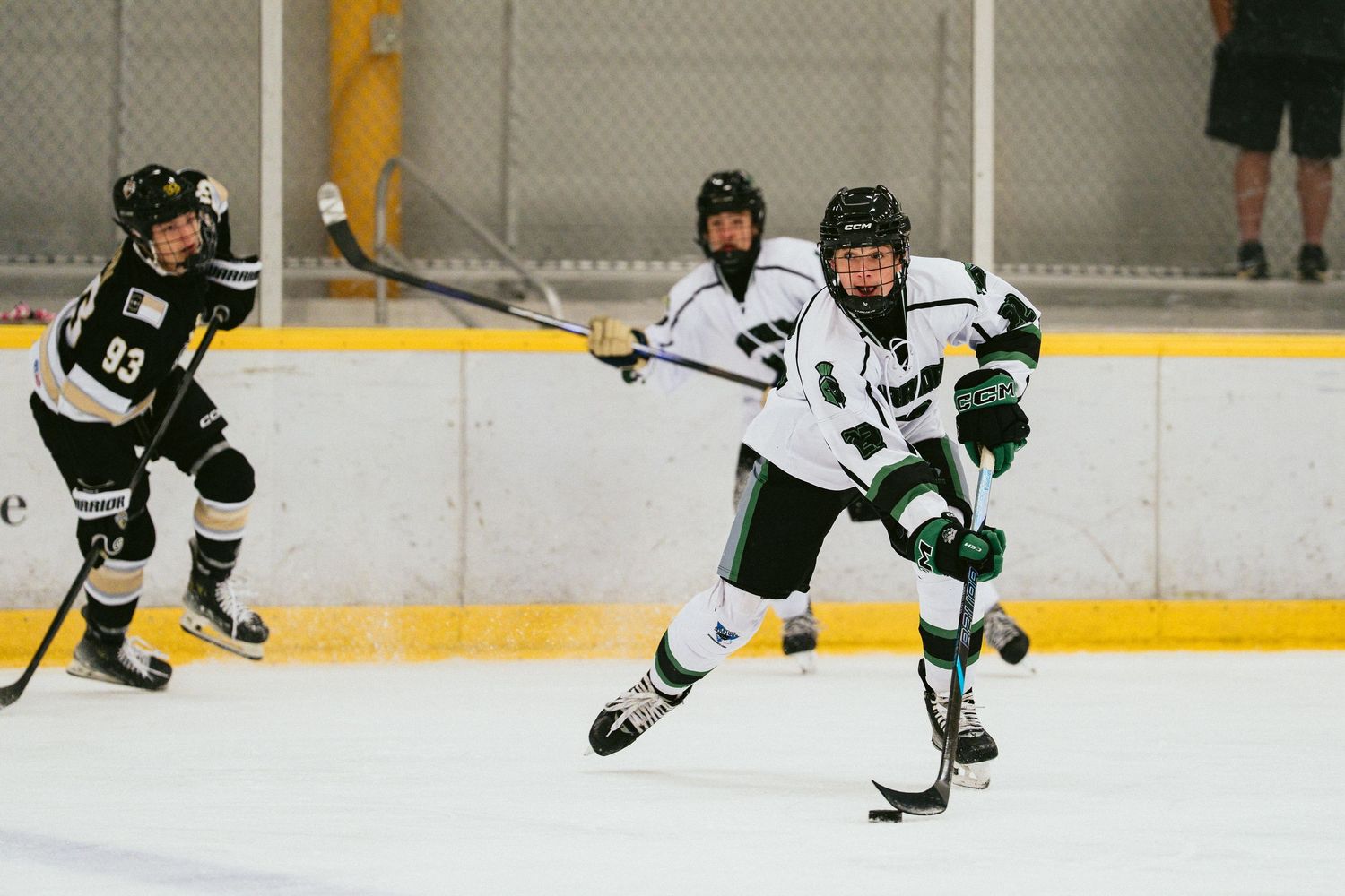 Youth ice hockey players compete on rink, player in white and green uniform skating with stick during game.