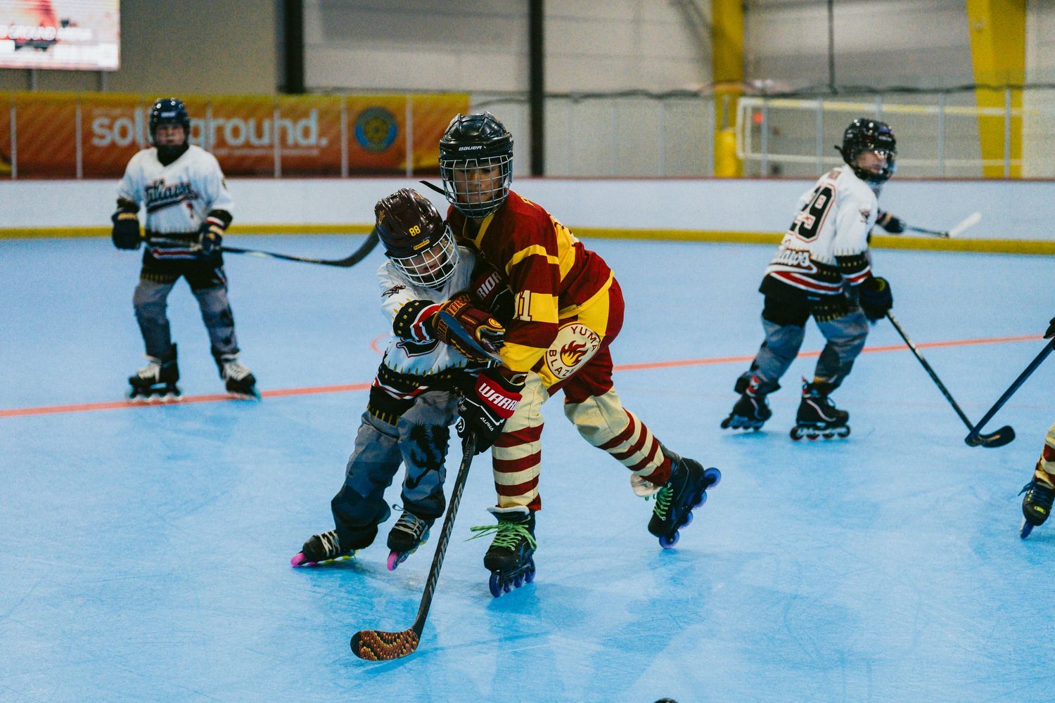 Kids playing inline roller hockey on a blue indoor rink, competing for the puck during a youth league game.