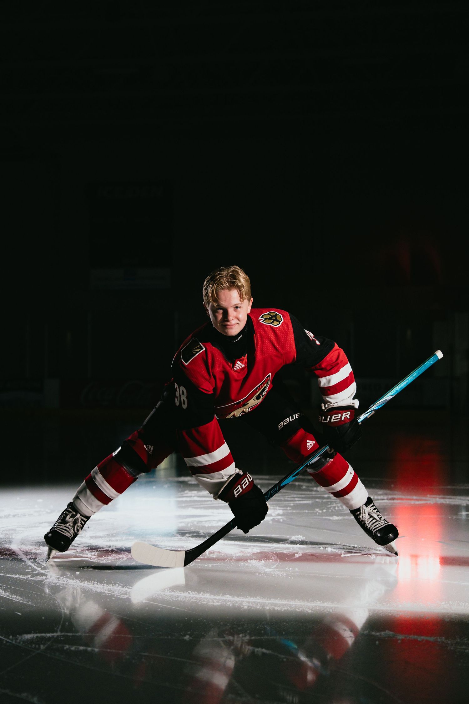 Hockey player in red uniform crouches on ice rink holding stick in dramatic dark arena lighting.