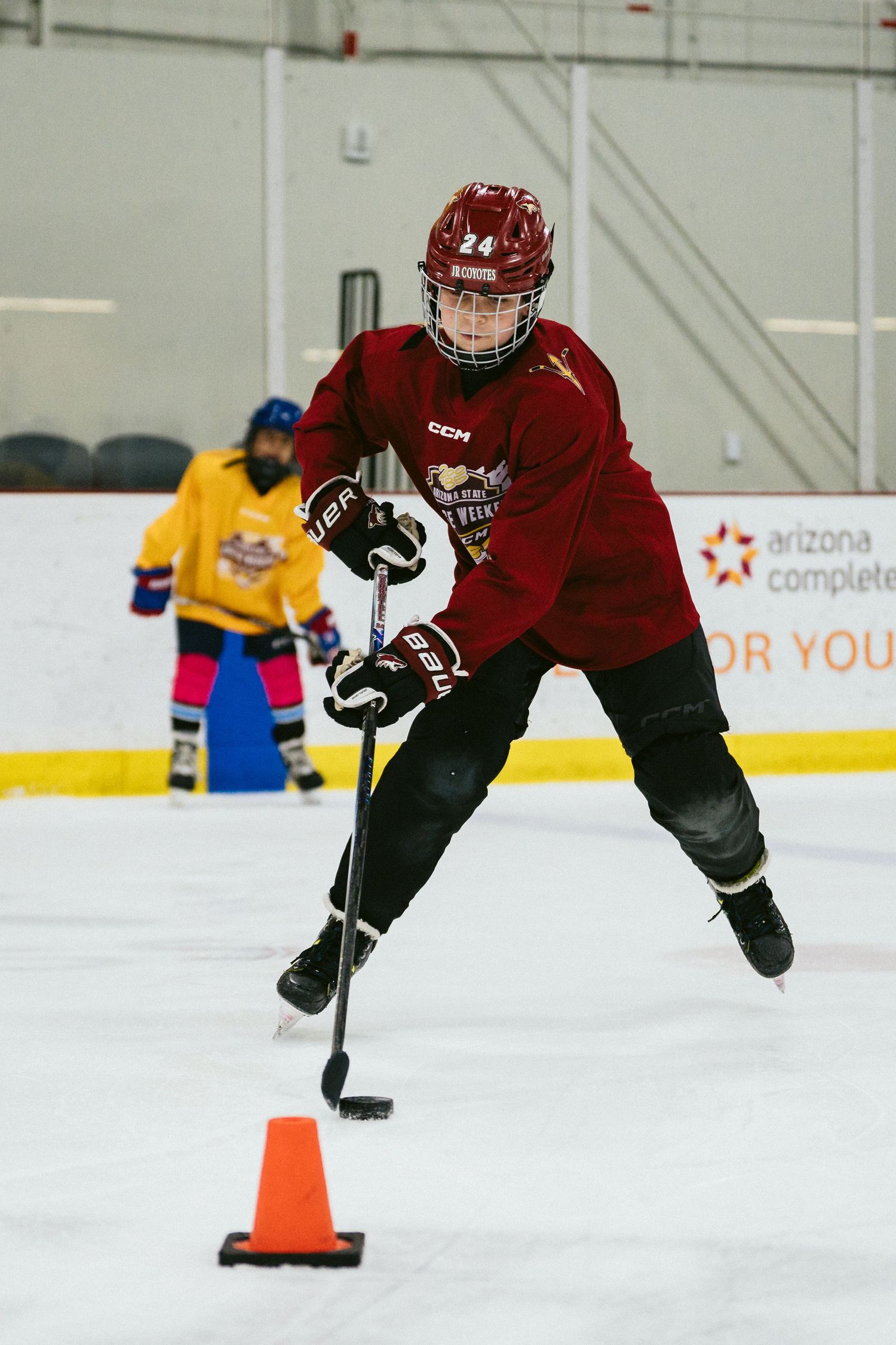 Youth hockey player in red jersey number 24 handles the puck around an orange cone during ice rink drills.