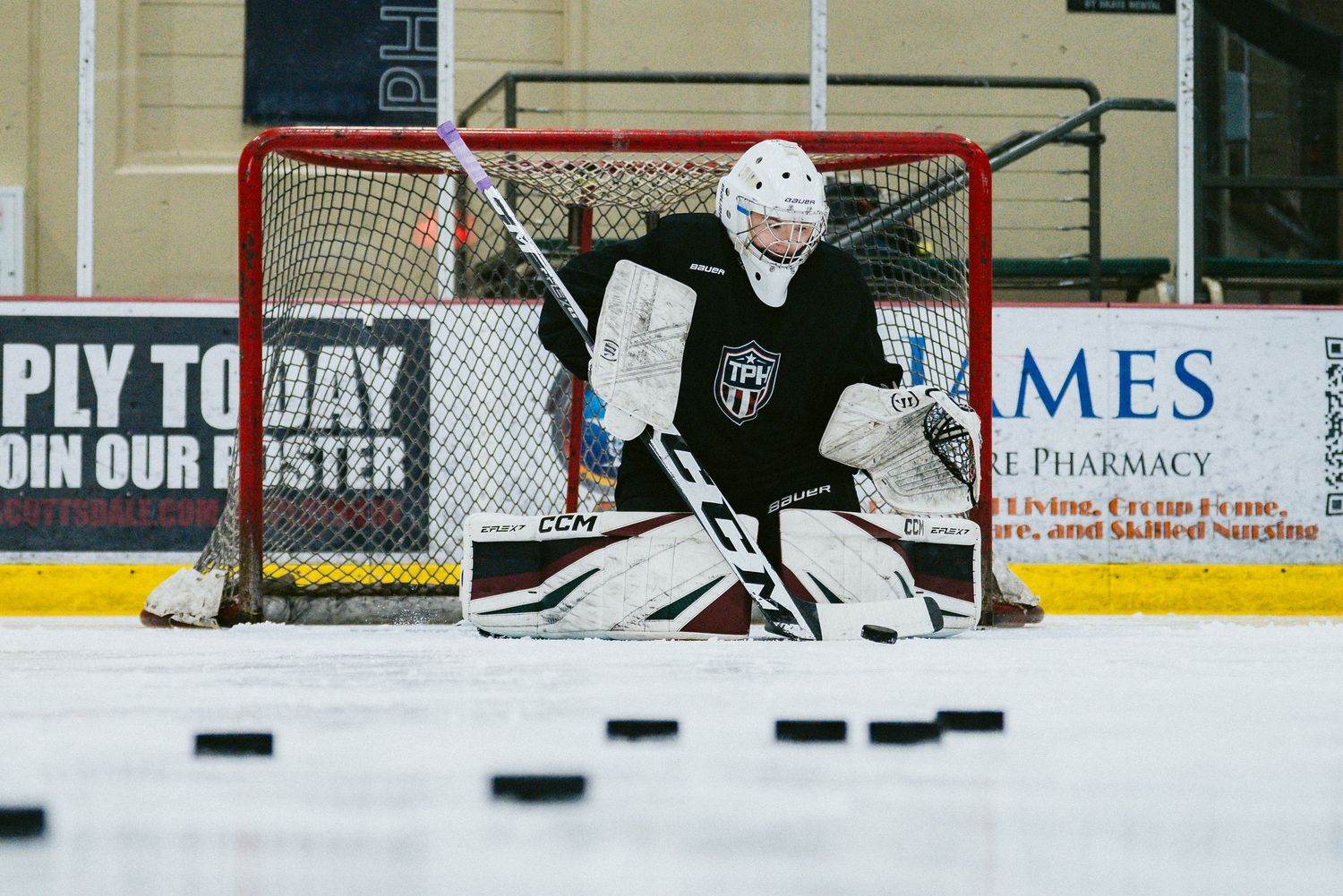 Hockey goalie in black jersey guards the net during practice, with multiple pucks on the ice rink.