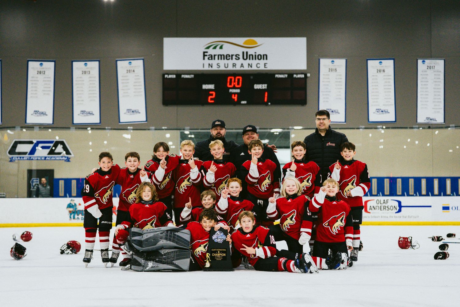 Youth hockey team in red jerseys poses for group photo on ice with coaches and trophy banners.