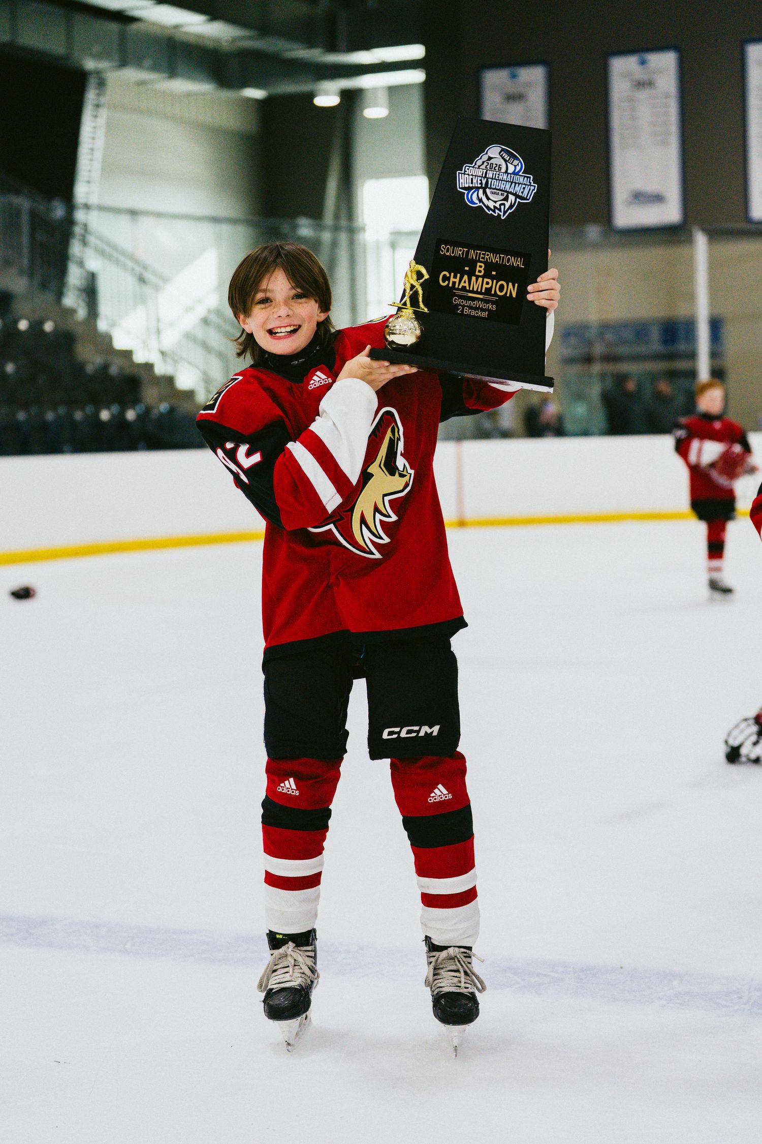 Young hockey player in red uniform showing off championship award on ice rink.
