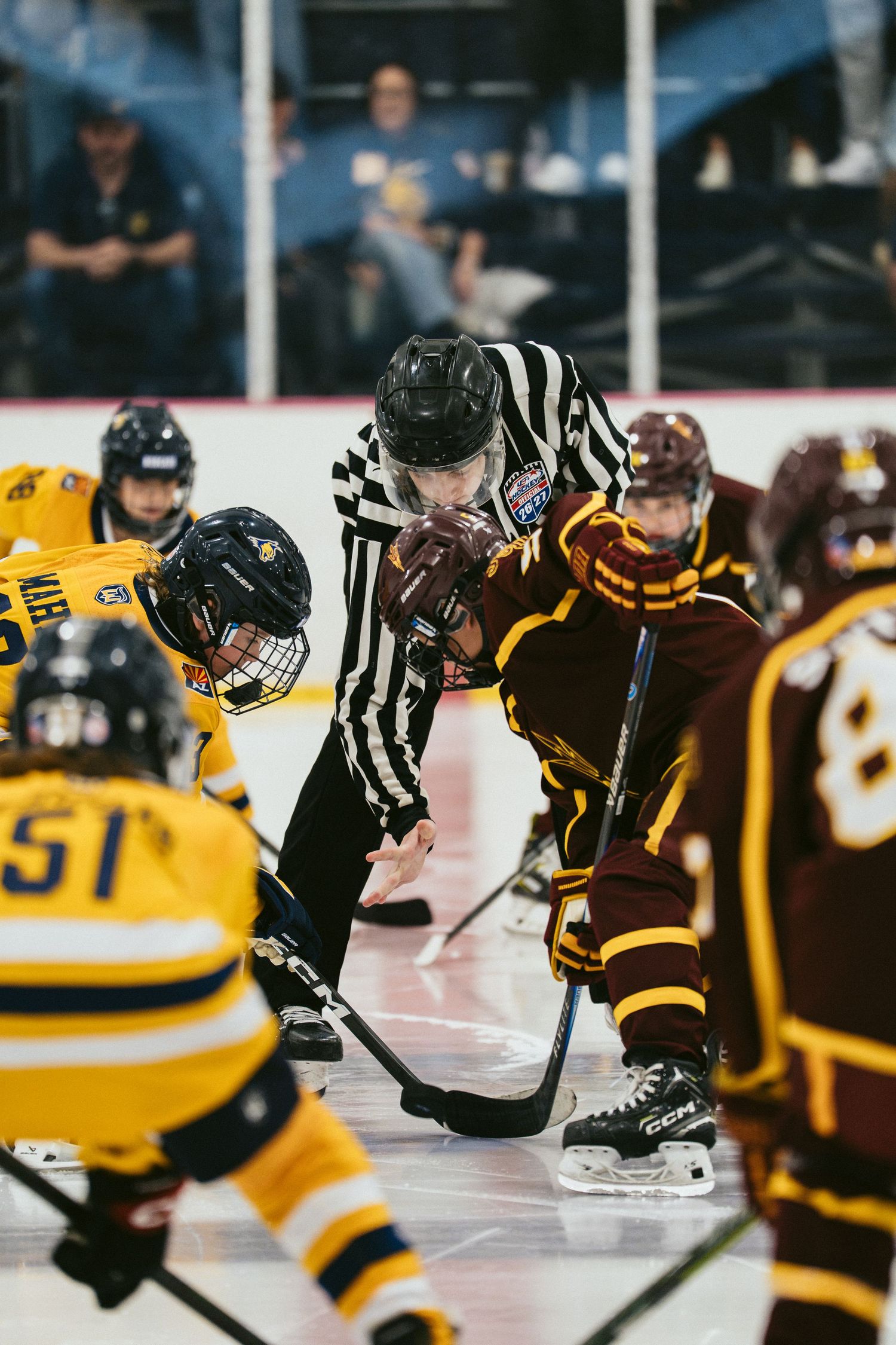Referee drops the puck at a face-off between maroon and gold youth hockey teams on an indoor ice rink.