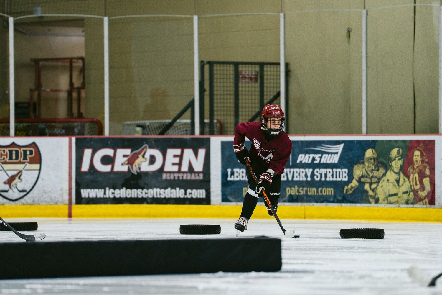 Youth hockey player in dark maroon jersey skates with puck at Ice Den Scottsdale indoor hockey rink.