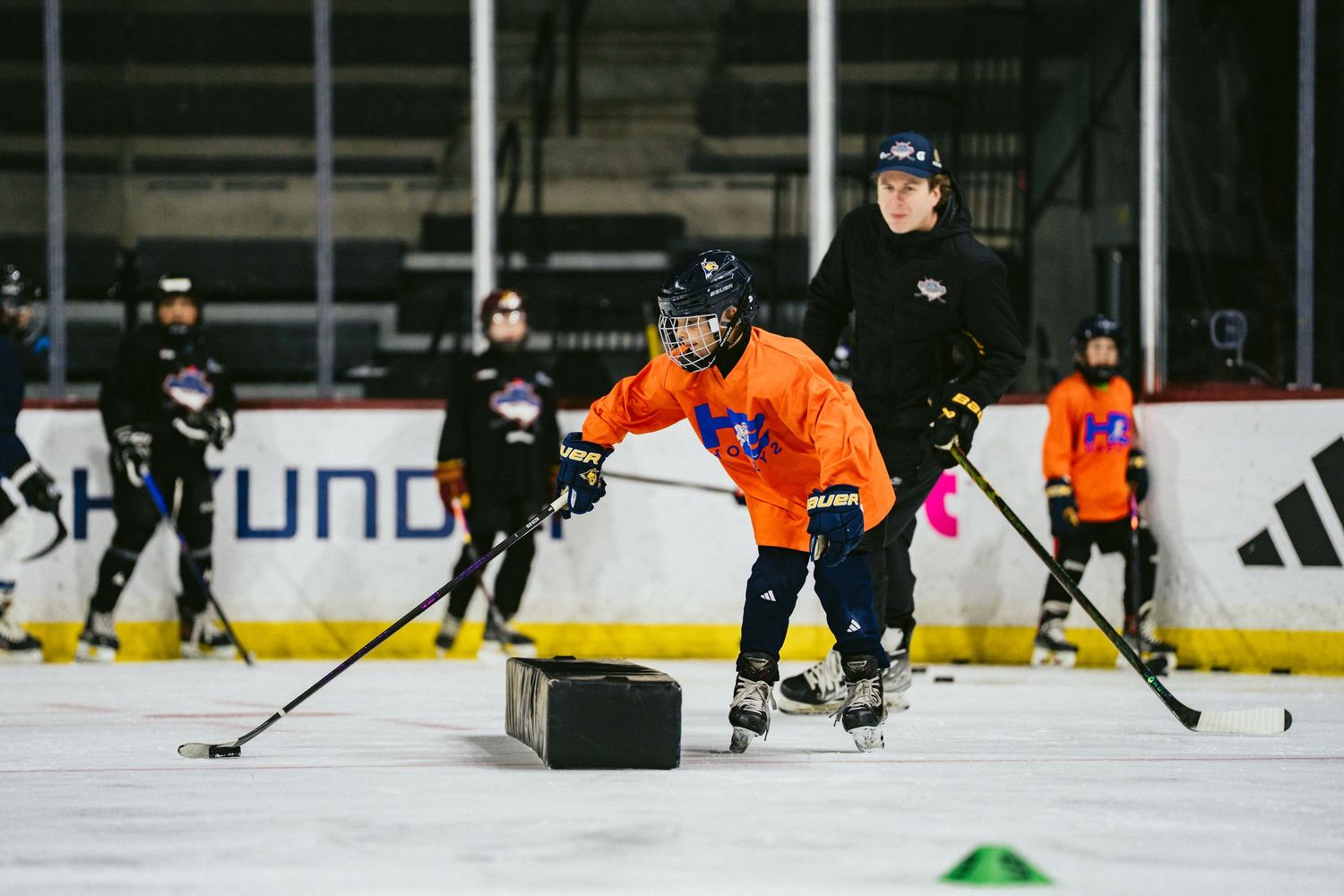 Young hockey player in orange jersey practicing skating drills with coach during ice clinic.
