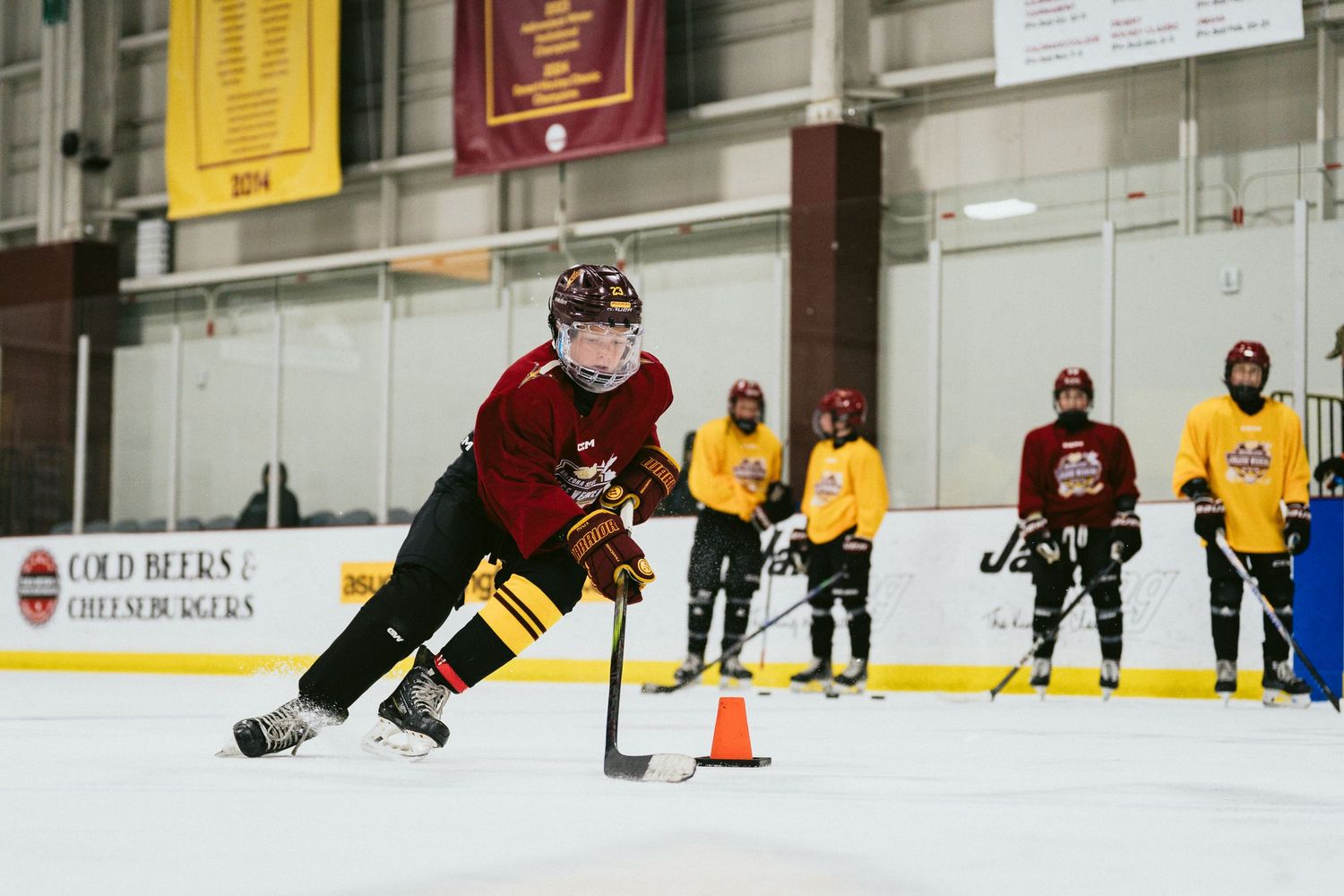 Youth hockey player dribbling puck around orange cone during ice skating drill with teammates watching.