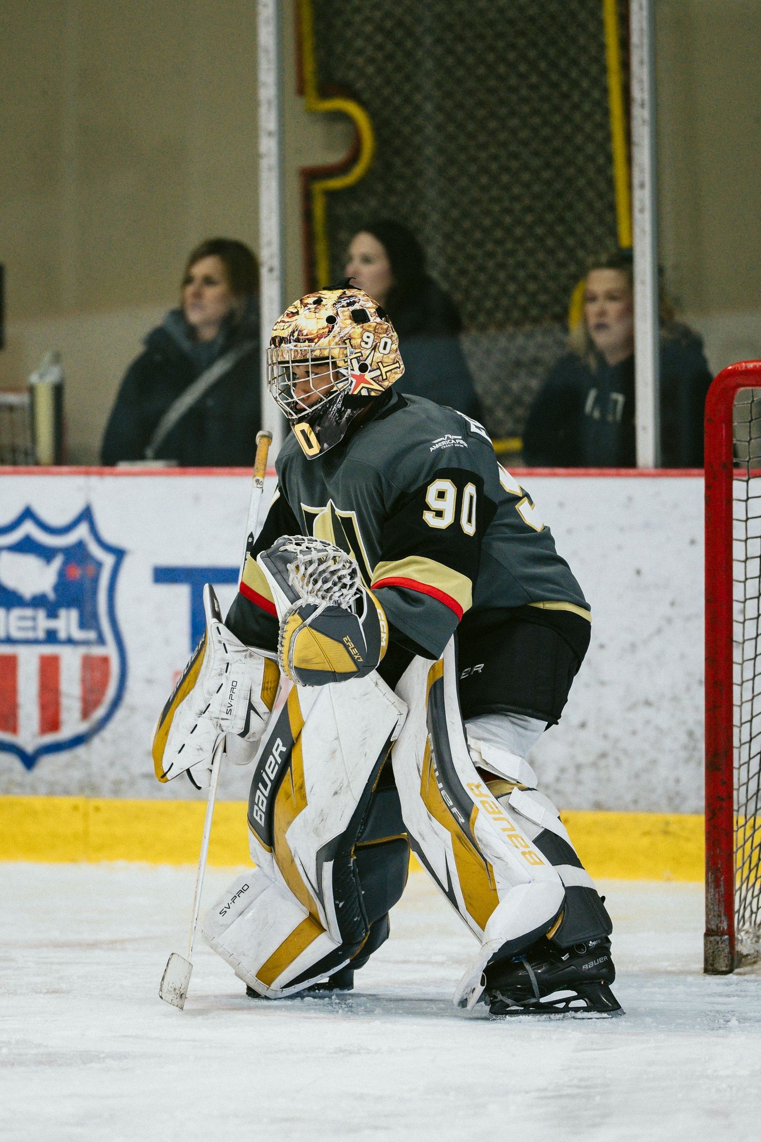 Hockey goalie in black and gold uniform crouches in defensive position guarding the net.