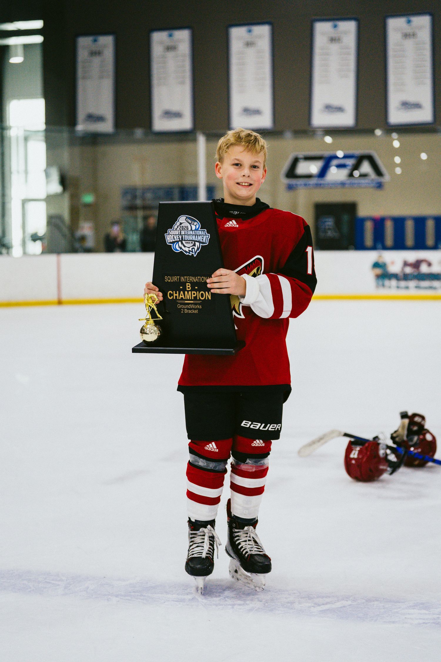 Youth hockey player in red uniform holds black award plaque on ice.