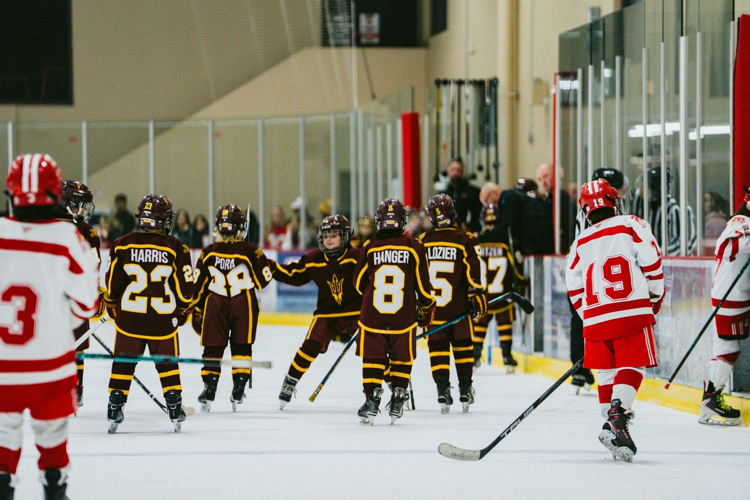 Youth hockey players in brown and red jerseys compete during an ice hockey game at an indoor rink.