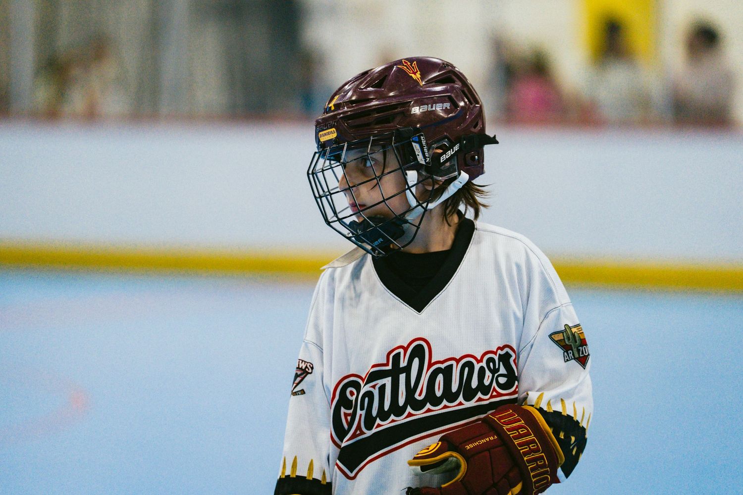 Youth hockey player in Outlaws jersey and maroon helmet stands ready during an indoor roller hockey game.