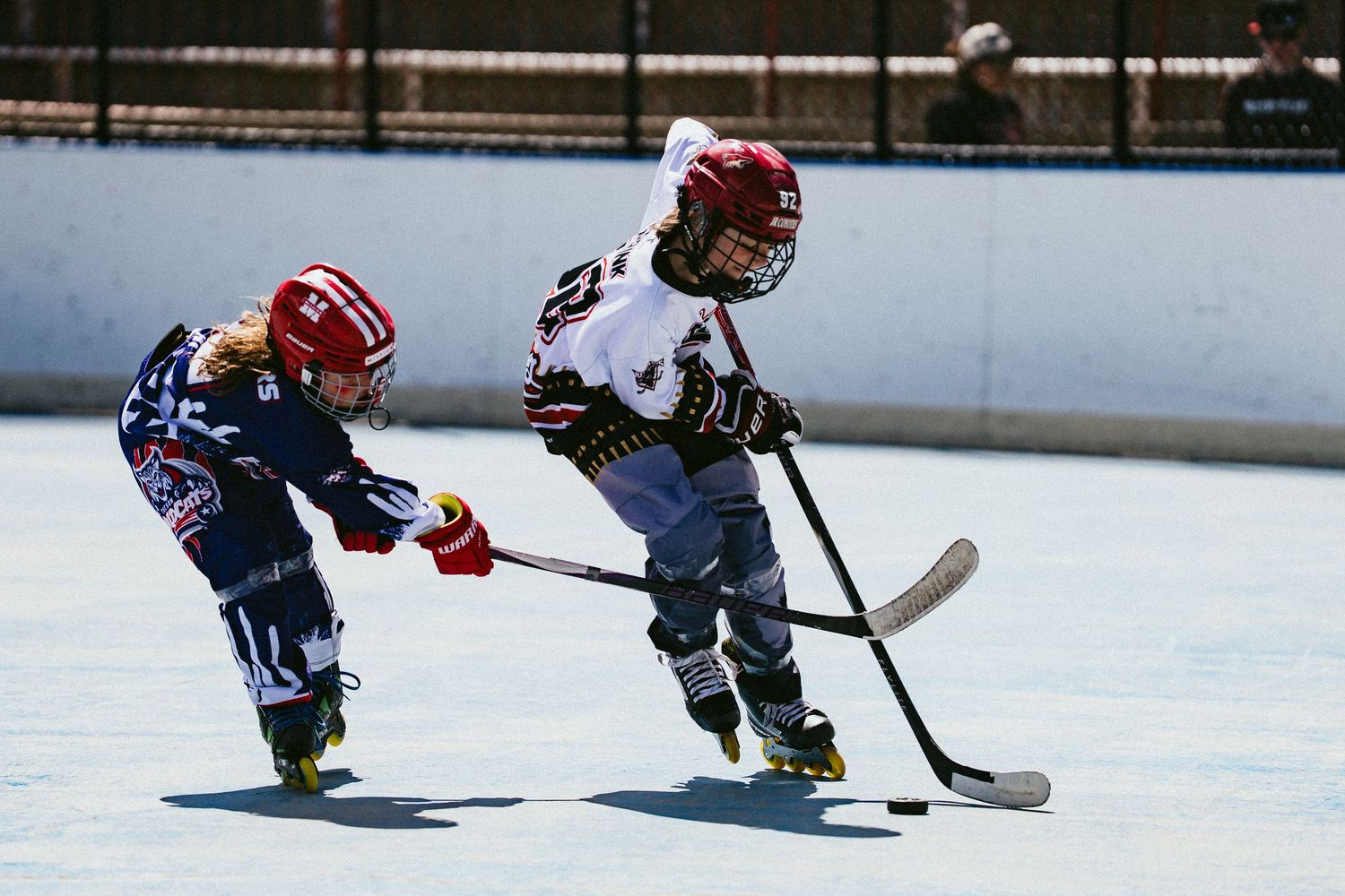 Two young kids in helmets compete for the puck during an outdoor roller hockey game.