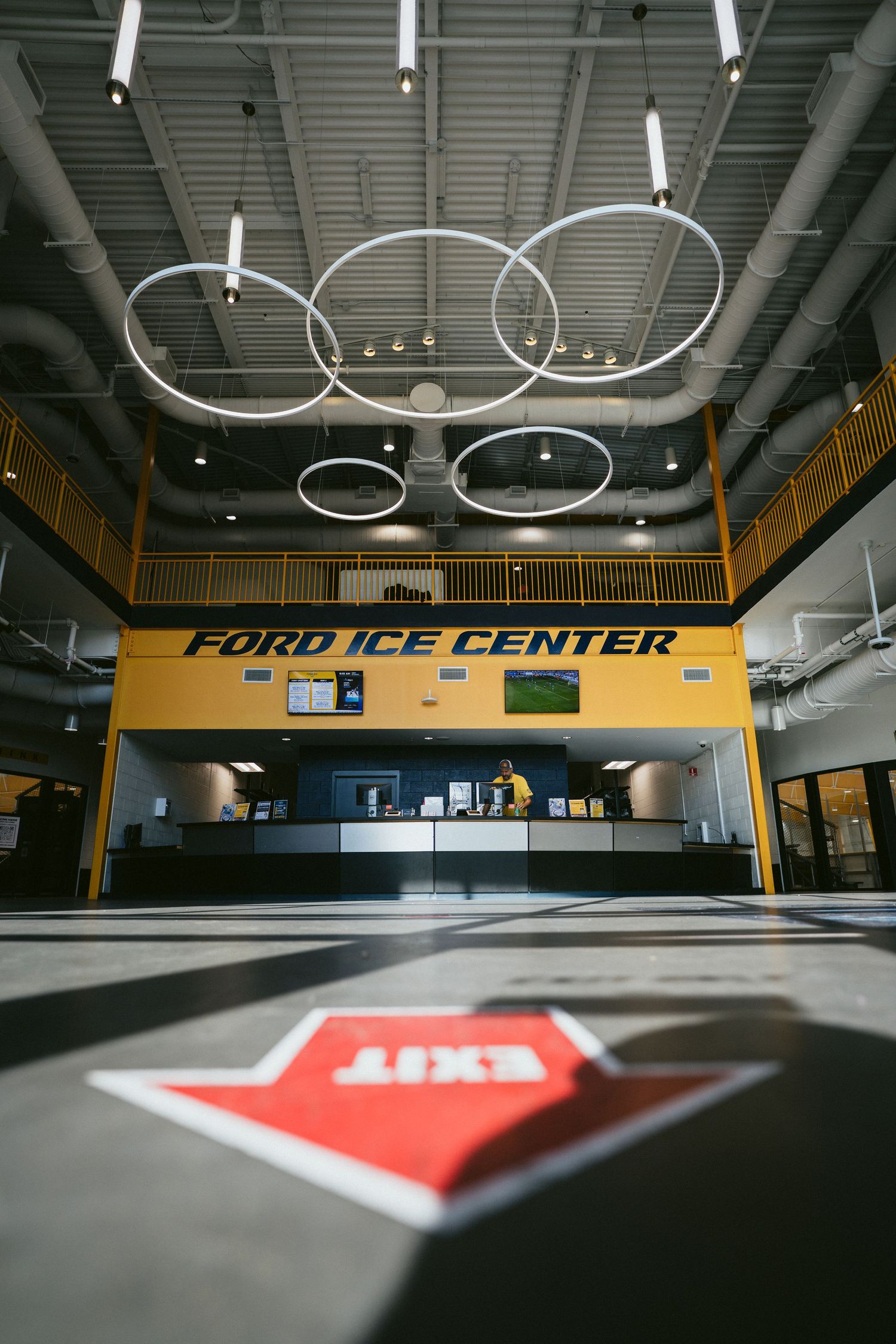 Modern Ford Ice Center lobby featuring circular ring lights, yellow signage, and a front desk with EXIT floor marking.
