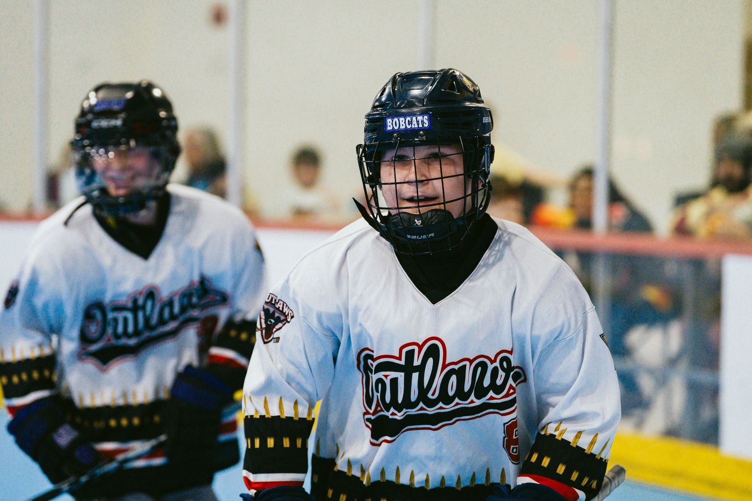 Two youth hockey players in Outlaws jerseys and helmets stand ready on an indoor roller hockey rink.