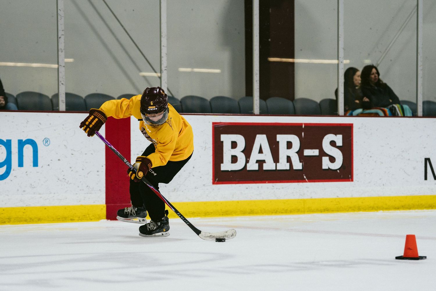Young hockey player in yellow jersey skates around an orange cone during an indoor ice hockey drill.