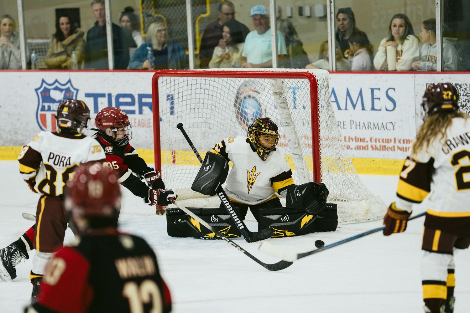 Women's ice hockey goalie makes a save during a competitive game while teammates defend the net on indoor ice rink.
