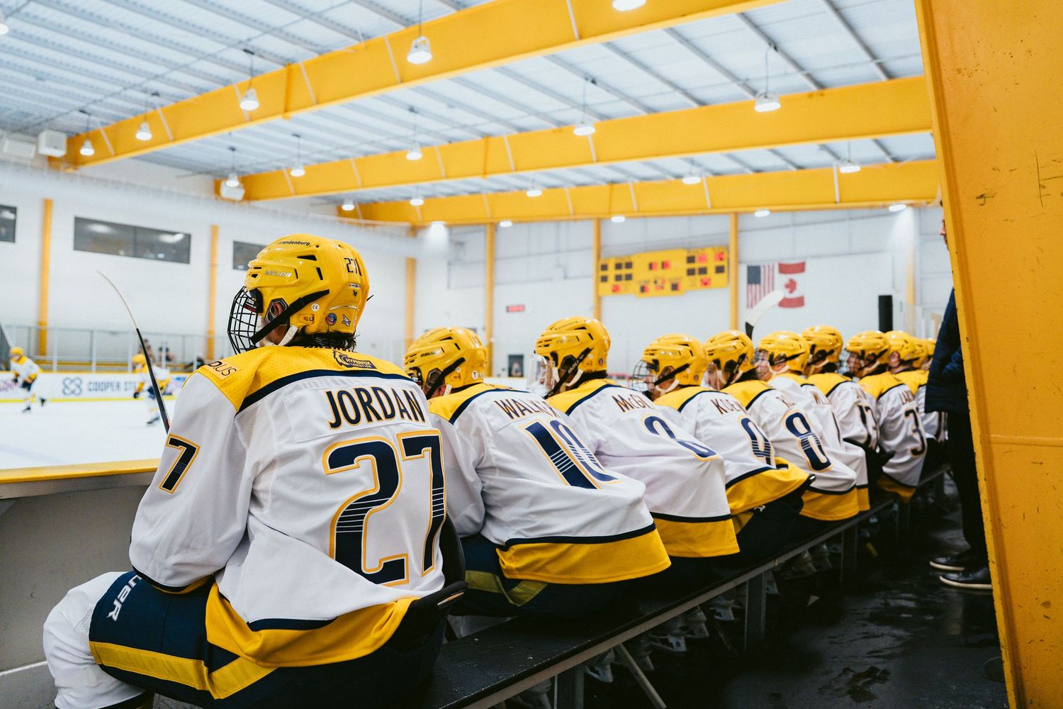Ice hockey team in yellow and white Jordan jerseys sit on bench during game at indoor rink.